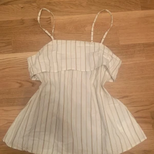 Vit randig linnetopp med volang - Säljer en söt vit linnetopp med tunna axelband och volang upptill. Toppen har ett rakt snitt och tunna vertikala ränder i beige. Perfekt för varma dagar!