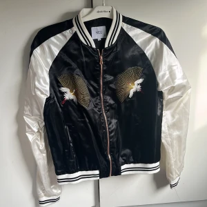 Svart och vit bomberjacka från lager 157 - Säljer en svart och vit bomberjacka från lager 157 med broderade tranor och solstrålar på framsidan. Jackan har dragkedja, randiga muddar och är tillverkad i glansigt material. Perfekt statement-plagg för dig som vill sticka ut.