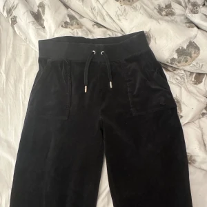 Svarta velourbyxor från Juicy Couture - Mysiga svarta velourbyxor från Juicy Couture med bred resår i midjan och snörning framtill. Byxorna har fickor både fram och bak, samt dekorativa knappar på bakfickorna. Perfekta för en avslappnad och trendig look.sönder vid fötterna