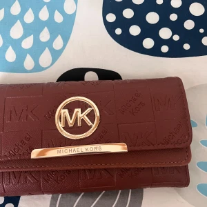 Brun och rosa plånbok från Michael Kors - Två stilrena plånböcker från Michael Kors, en i brun och en i stark rosa. Båda har guldiga MK-detaljer och präglad logga. Plånböckerna har flera kortfack, fönster för ID och plats för sedlar. Perfekt accessoar för dig som vill ha både stil och funktion.