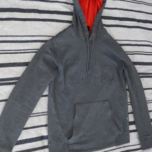 Grå hoodie från Hugo Boss med röd huva - Säljer en stilren grå hoodie från Hugo Boss med broderad logga på bröstet. Insidan av huvan är röd för en snygg kontrast. Tröjan har känguruficka och dragsko i huvan. Perfekt för en chill och trendig look.