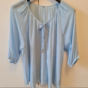 Ljusblå blus med knyt och fjäderdetaljer från KappAhl - Säljer en ljusblå blus med trekvartsärm och knyt framtill med snygga guldfärgade fjäderdetaljer från KappAhl. Blusen har en lös passform och är perfekt för vår och sommar. Storlek 46 (48). Etiket klippt. Kan hämtas i Vallentuna.