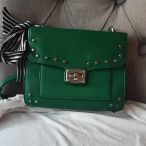 Grön axelväska med nitar och rosett - Snygg grön axelväska med silverfärgade nitar och ett dekorativt spänne framtill. Väskan har en svartvit randig rosett på sidan och justerbar axelrem. Perfekt för att addera färg till din outfit.