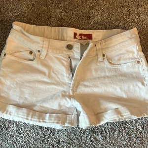 Vita jeansshorts från &NOW: - Snygga vita jeansshorts från &NOW: med klassisk femficksdesign och uppvikta benslut. Shortsen har knapp och dragkedja framtill samt bälteshällor. Perfekta för varma dagar och enkel att matcha med olika toppar.