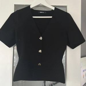 Svart v-ringad topp med dekorativa knappar från Gina Tricot - Snygg svart topp från Gina Tricot med v-ringning och tre unika silvriga knappar framtill.Använder knappast denna tröja längre men inget fel på den och sitter bra. Tröjan är kort i sin storlek då jag brukar ha xs men ville ha den lite längre. Original pris 400kr säljer för 169kr. Använt en gång på jul.