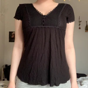 babydoll topp  - Svart blus med v-ringning och dekorativa knappar framtill. Spetsdetaljer längs halsringningen och ärmarna. Korta ärmar och lätt utställd passform. Perfekt för en stilren look.