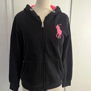 Zip up hoodie från Ralph Lauren storlek L (barnstorlek) - Marinblå  zip-up hoodie från Ralph Lauren med dragkedja och rosa innerfoder i huvan. Stor rosa logga på bröstet och siffra på ärmen. Praktiska fickor framtill och ribbade muddar. Är i storlek L i barn. Den har en liten fläck vid fickan men den syns knappt.