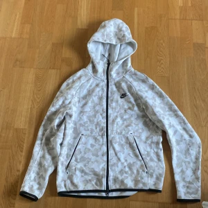Vit camomönstrad hoodie från Nike - Säljer en vit hoodie från Nike med ljusbeige camomönster. Jackan har huva, hel dragkedja och två fickor med dragkedja. Svarta detaljer längs dragkedja och ärmslut. Perfekt för en sportig och cool stil.