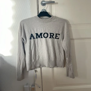 Beige tröja med text från Gina Tricot - Säljer en beige tröja från Gina Tricot med svart texten 'AMORE' framtill. Tröjan har rund halsringning och långa ärmar. Perfekt för en avslappnad och stilren look.