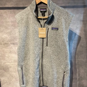 Patagonia Väst - Säljer en grå fleeceväst från Patagonia med hel dragkedja framtill och två sidofickor med dragkedja. Västen har hög krage och klassisk logga på bröstet. Perfekt för lager-på-lager och enkel att matcha. 