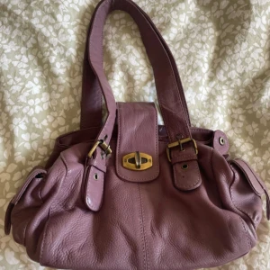 Lila handväska från Depeche - Snygg lila handväska från Depeche i skinn. Väskan har praktiska fickor med dragkedja och dekorativa spännen. Perfekt för att hålla ordning på dina saker med stil.