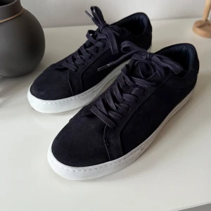 Marinblåa sneakers från Les Deux - Snygga marinblåa sneakers i mocka från Les Deux med vita sulor och snörning. Diskreta broderade detaljer i rött och blått på hälen ger en stilren touch. Perfekta för en avslappnad look. Storlek 42. De är i mycket bra skick. 