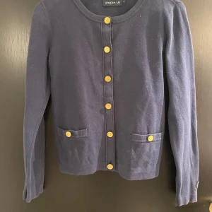 Mörkblå kofta från Stockh LM - Säljer en stilren mörkblå kofta från Stockh LM med guldknappar framtill och två fickor. Koftan har långa ärmar och en rund halsringning. Perfekt för en elegant look.