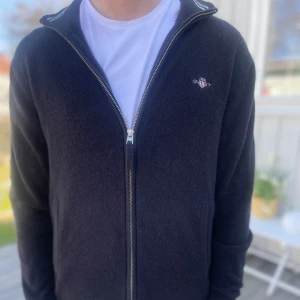 Svart kofta från Gant - Säljer en stilren svart kofta från Gant med dragkedja och broderad logga på bröstet. Perfekt för både vardag och mer uppklädda tillfällen. Koftan har långa ärmar och en bekväm passform.