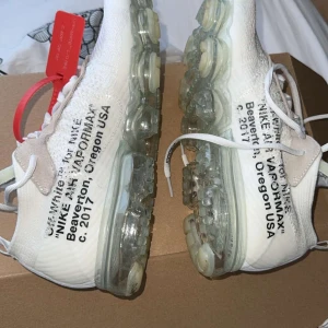 Nike Air Vapormax Off-White - Säljer ett par exklusiva Nike Air Vapormax i samarbete med Off-White. Skorna är vita med transparent sula och har den ikoniska Off-White texten på sidan. De har snörning och en röd tagg som detalj. Perfekta för den som vill ha något unikt och stilrent.