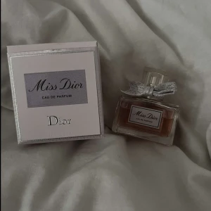 Miss Dior Eau de Parfum - Elegant och tidlös Miss Dior Eau de Parfum från Dior. Flaskan har en stilren design med en söt rosett på korken. Doften är blommig och feminin, perfekt för den som älskar en klassisk och sofistikerad parfym.