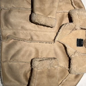 Beige teddyjacka med fuskpäls - Säljer en beige teddyjacka från Zara med mjukt foder och krage i fuskpäls. Jackan har knappar framtill och långa ärmar med pälsdetaljer vid ärmslut och krage. Perfekt för kyliga dagar!