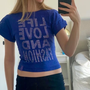 Lila/Blå croppad t-shirt med text - Snygg blå croppad t-shirt med texten 'LIFE LOVE AND FASHION' i glittriga stenar framtill. T-shirten har korta ärmar och en tight passform, perfekt för en trendig look. Från kappahls barnavdelning från början men sitter perfekt på mig som är xs