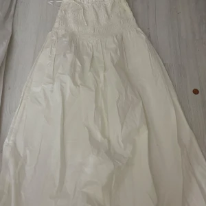 Vit långklänning med smockdetaljer - Oanvänd elegant vit långklänning med smockdetaljer upptill och tunna axelband. Perfekt för sommarens festligheter. Klänningen har en luftig passform och är tillverkad i ett lätt material som ger en skön känsla.