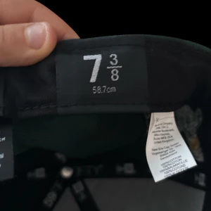 Grön keps med broderad logotyp - Snygg grön keps från New Era med broderad logotyp framtill. Kepsen har en klassisk design med en vit skärm och MLB-logotypen baktill. Perfekt för sportiga tillfällen eller som en stilren accessoar.