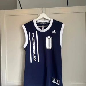 Blå ärmlös baskettröja från Adidas - Säljer en blå ärmlös baskettröja från Adidas med texten 'THUNDER' och nummer 0 på framsidan. Baksidan har namnet 'WESTBROOK' och nummer 0. Perfekt för basketfans! 🏀