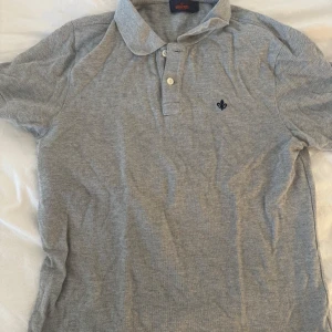 Grå pikétröja från Morris - Säljer en klassisk grå pikétröja från Morris med krage och två knappar framtill. Tröjan har korta ärmar och en liten broderad logga på bröstet. Perfekt för en stilren och avslappnad look.