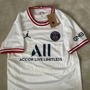 PSG vit fotbollströja Jordan x Paris Saint-Germain - Säljer en vit PSG fotbollströja med Jordan-logga och Paris Saint-Germain-emblem. Tröjan har korta ärmar, röd, blå och vit randig krage och ärmslut. Stora tryck på bröstet och sponsorlogga. Perfekt för fotbollsfans!