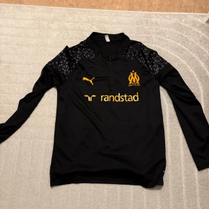 marseille tröja - Säljer en svart långärmad tröja från Puma med gul logga och text. Tröjan har ett diskret mönster på axlarna och en dragkedja vid halsen. Perfekt för sportiga aktiviteter eller som en stilren vardagströja. Helt Ny!