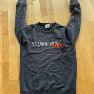 Svart sweatshirt från Dondup - Säljer en stilren svart sweatshirt från Dondup med tryck i vitt och rött på framsidan. Tröjan har långa ärmar och en rund halsringning. Perfekt för en avslappnad look.