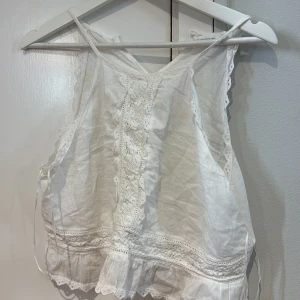 Vit spetsdetaljerad linnetopp från Primark - Söt vit linnetopp från Primark med tunna axelband och fina spetsdetaljer framtill och längs kanterna. Toppen har broderade inslag och volang nertill, perfekt för varma dagar.passar mig som har S fats den är 42