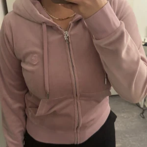 Rosa velour hoodie från Juicy Couture - Säljer en klassisk rosa hoodie i mjuk velour från Juicy Couture. Tröjan har dragkedja, huva med snören och fickor framtill. Perfekt för en avslappnad och trendig look. Ikonisk JC-logga broderad på bröstet.