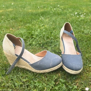 Blåa espadriller med kilklack från Bata - Säljer ett par blåa espadriller från Bata med kilklack och justerbar rem runt vristen. Skorna har ovandel i tyg och flätad sula i repimitation för en somrig look. Perfekta för dig som vill ha både stil och komfort. Storlek 37 
