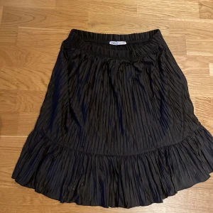 Svart plisserad kjol från Zara - Säljer en svart plisserad kjol från Zara med bred resår i midjan och volang nertill. Kjolen har en luftig känsla och är perfekt för dig som gillar en klassisk men trendig stil.50:-