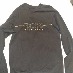Svart tröja från Hugo Boss - Snygg svart tröja från Hugo Boss med deras ikoniska logga i vitt och gult på bröstet. Tröjan har långa ärmar och en klassisk rund halsringning. 