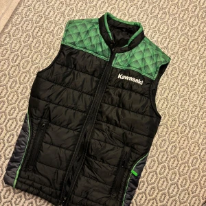 Svart och grön dunväst från Kawasaki - Snygg svart dunväst från Kawasaki med gröna detaljer och quiltad ovandel. Västen har dragkedja framtill, två fickor med dragkedja och stor Kawasaki-logga både fram och bak. 