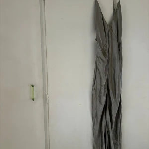 BALKLÄNNING Ljusgrå långklänning med v-ringning silke  - Säljer en elegant ljusgrå långklänning med smala axelband och djup v-ringning. Klänningen har omlott-effekt framtill och är tillverkad i ett glansigt material som ger en lyxig känsla. Perfekt för speciella tillfällen! Materialet är i Silke. Slits med. Prislapp på!