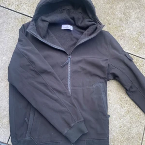 Svart jacka från Stone Island - Säljer en svart jacka från Stone Island med huva och dragkedja. Jackan har långa ärmar och är tillverkad i ett syntetmaterial med fleecefoder för extra värme. Perfekt för kyligare dagar.