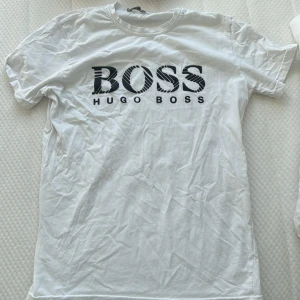 Vit t-shirt från Hugo Boss - Säljer en stilren vit t-shirt från Hugo Boss med stor logga i svart på bröstet. Perfekt för en casual look. T-shirten har en klassisk passform och är kortärmad.