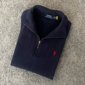 Mörkblå tröja med dragkedja från Ralph Lauren - Säljer en stilren mörkblå tröja från Ralph Lauren med halv dragkedja och det klassiska röda logotypbroderiet på bröstet. Perfekt för en avslappnad men snygg look. Tröjan har långa ärmar och är tillverkad i ett mjukt material.