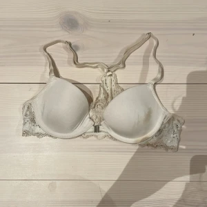 Vit spets-bh från Adore - Säljer en vit bh från Adore med vadderade kupor och fina spetsdetaljer på sidorna och mellan kuporna. Det har en fläck på ena sidan