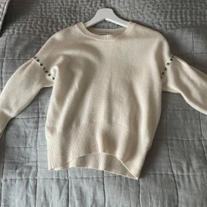 Vit stickad tröja med nitar - Säljer en vit stickad tröja med rund hals och långa ärmar. Tröjan har dekorativa nitar längs axlarna och bred ribbad mudd vid midjan och ärmsluten. Perfekt för en stilren och trendig look.