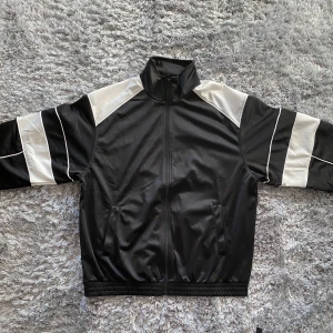 Jacka - Snygg svart baggy jacka med vita detaljer och dragkedja. Jackan har långa ärmar och en cool look, perfekt för en avslappnad stil. Den har en hög krage och elastiska muddar för en bekväm passform.