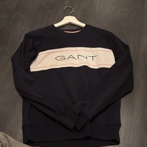 Mörkblå sweatshirt från GANT - Säljer en mörkblå sweatshirt från GANT med en bred vit panel och logga framtill. Tröjan har rund halsringning och långa ärmar. Väl använd men ingenting som syns.