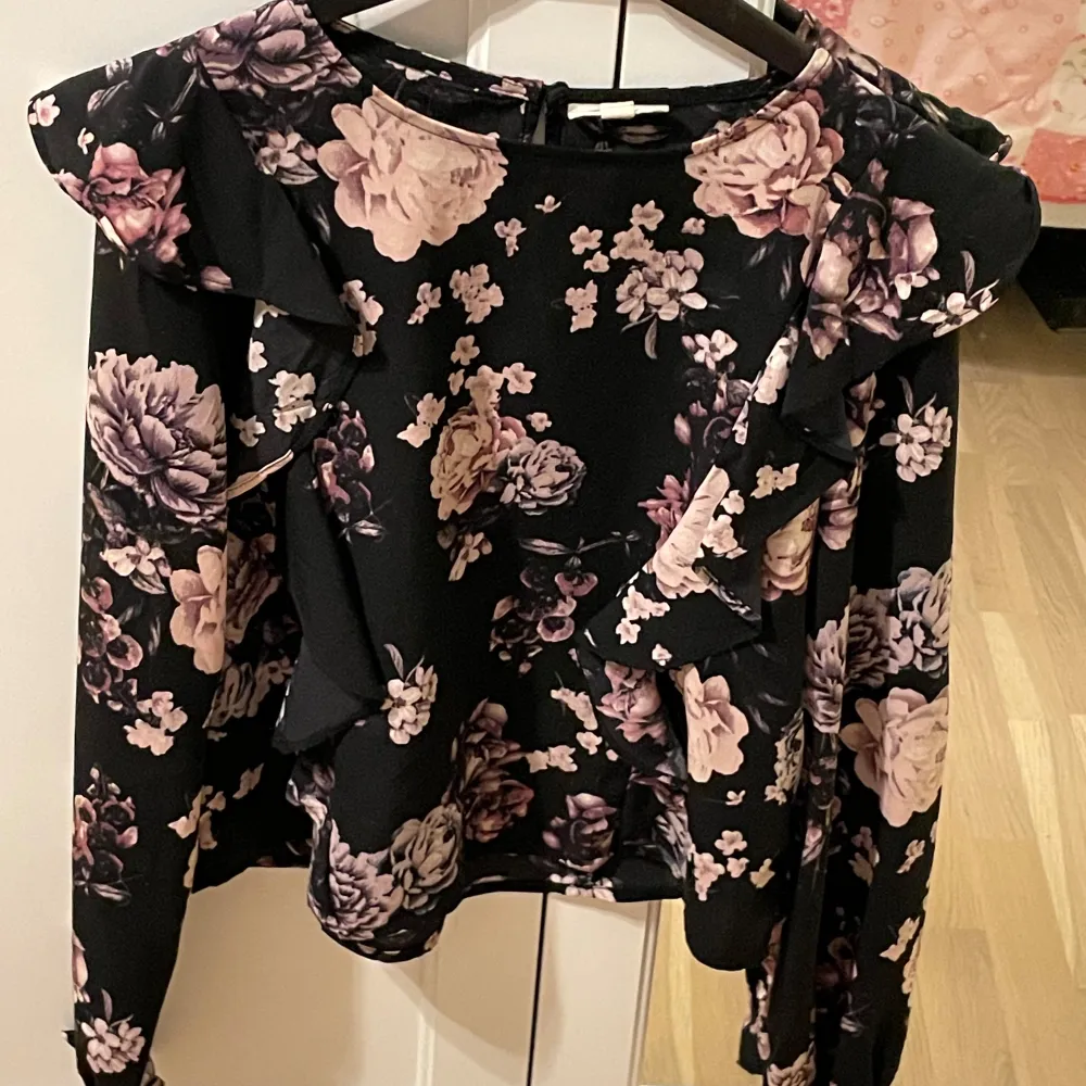 Supersnygg svart blus från KappAhl med lila och rosa blommor. Blusen har långa ärmar och volangdetaljer vid axlarna och framtill. Perfekt för dig som vill sticka ut lite extra med ett romantiskt mönster!. Puserot.