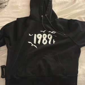 1989 taylor Swift - Svart hoodie med vit text '1989' och fågelmotiv på bröstet. Tröjan har huva med dragsko och en stor magficka framtill. Perfekt för en avslappnad stil. Taylor Swift