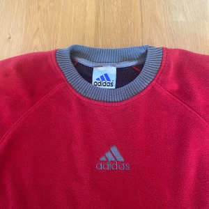 Röd fleece-tröja från Adidas - Säljer en röd fleece-tröja från Adidas med grå detaljer på sidorna, axlarna och kragen. Tröjan har rund halsringning och Adidas-logga broderad framtill. 