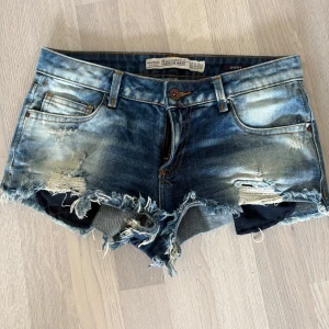 Blå korta jeansshorts från Zara - Säljer ett par korta blå jeansshorts från Zara med slitna detaljer och fransar. De har en knapp och dragkedja framtill samt klassiska femficksdesign. Perfekta för en avslappnad sommarlook. 
