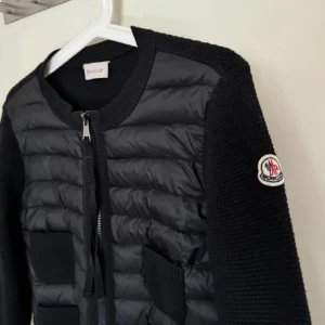 Moncler cardigan dam - Snygg svart quiltad jacka från Moncler med dragkedja framtill. Jackan har långa ärmar och en rund halsringning. Perfekt för kyligare dagar med sin stilrena design och bekväma passform, säljer min flickväns cardigan, storlek XS