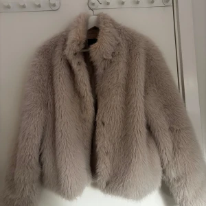 Beige pälsjacka med krage - Säljer en fluffig beige pälsjacka med lång ärm och hög krage. Jackan har en klassisk look och är perfekt för kyligare dagar. Stängs framtill med dolda knappar. Supermjuk och trendig modell!