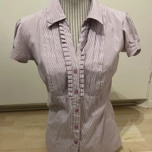 Röd- och vitrandig blus med volangdetalj - Säljer en söt blus med röda och vita smala ränder, korta puffärmar och volangdetaljer längs knappraden. Skjortkrage och knappar framtill. Omstydd från L till M/S. Hör gärna av er vid frågor. 